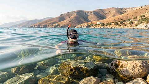 Apneista nadando en superficie sobre un fondo rocoso somero, agua turquesa clara y costa seca al fondo